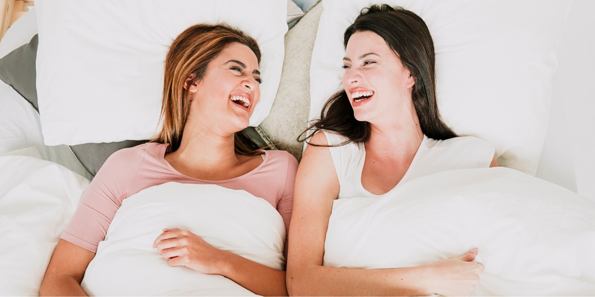 2 laughing women lying in bed - My first time with another woman